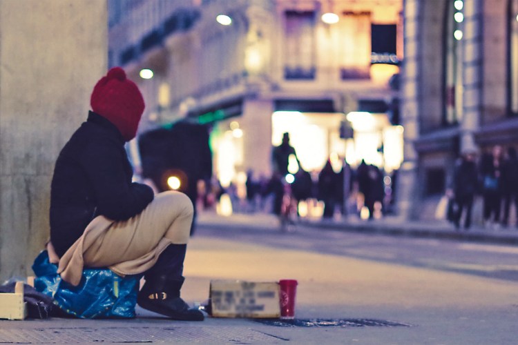 dressed in winter clothes, person sits on street as people shop at nearby lit-up storefronts.